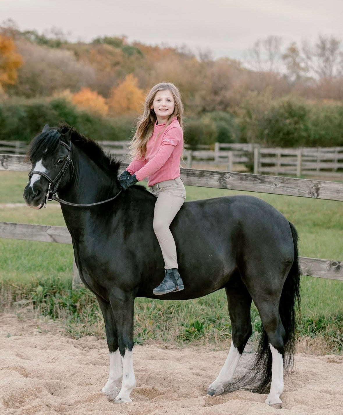 Young girl riding a black horse in a pink riding top, with trees in the background
