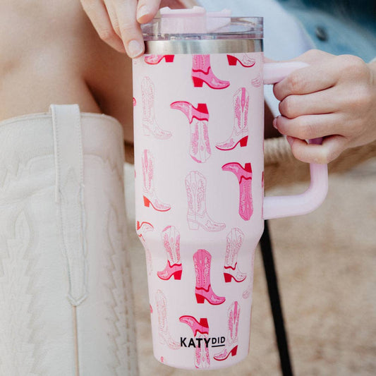 A pair of light pink western boots-themed tumbler cup with handle, featuring a stainless steel interior and a screw-on plastic lid.