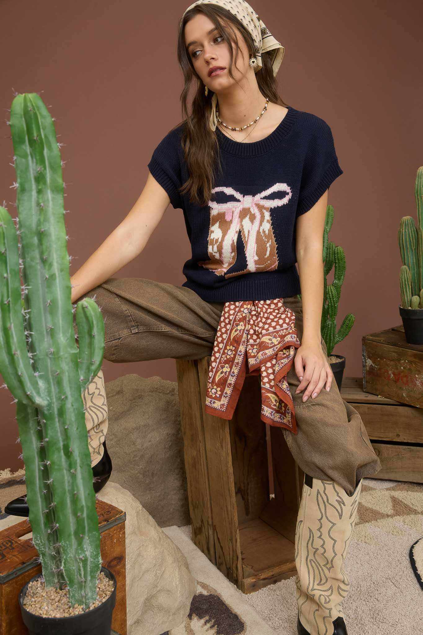 Woman sitting in a room with cacti and wooden furniture, wearing a dark blue sweater with a  cowgirl boot and bow design.