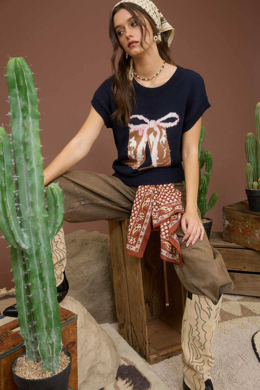 Woman sitting in a room with cacti and wooden furniture, wearing a dark blue sweater with a  cowgirl boot and bow design.