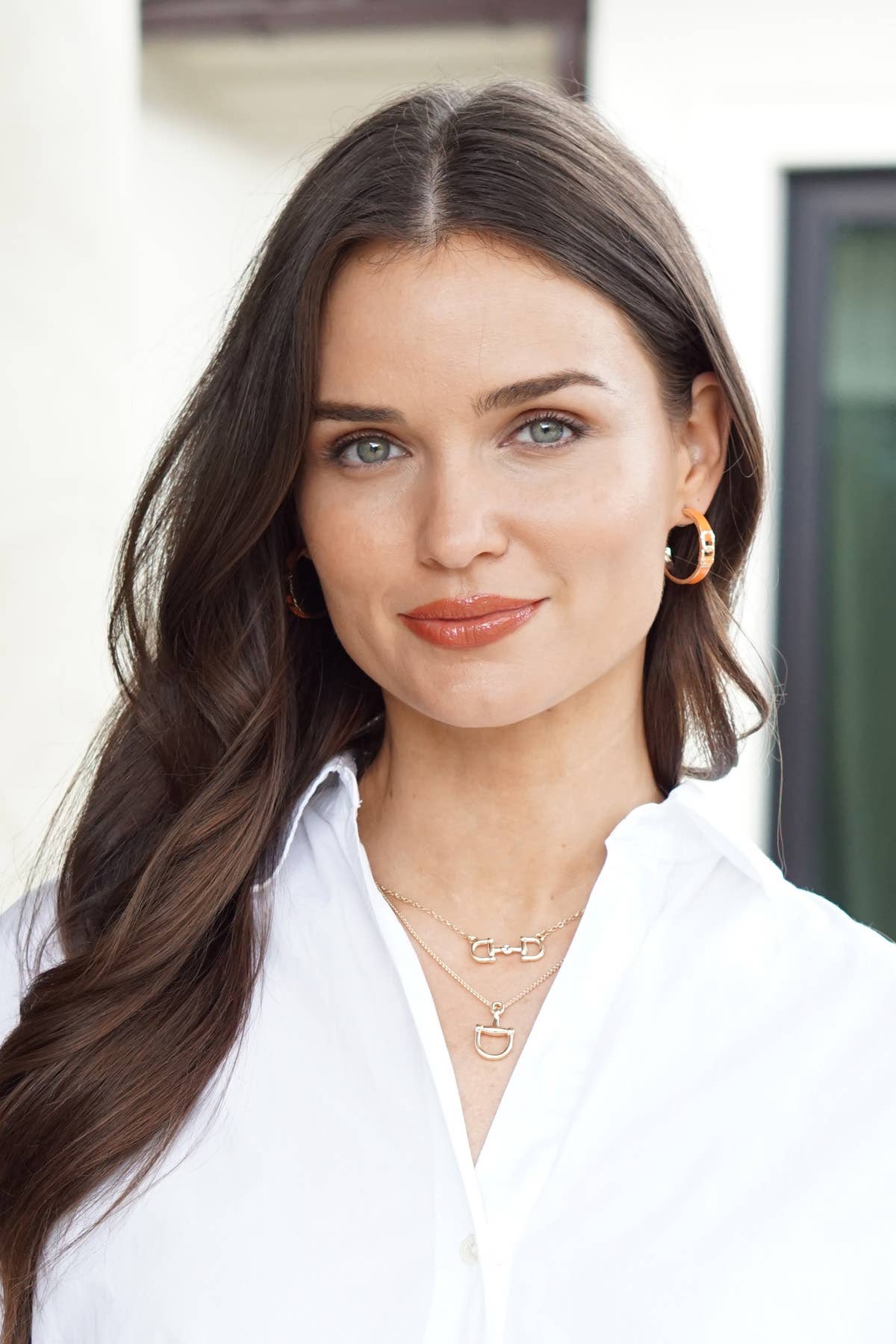 Woman with long brown hair wearing a white shirt and gold equestrian themed jewelry.