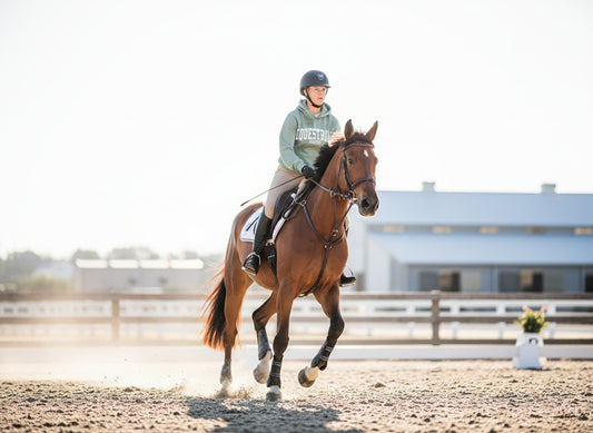 Person riding a horse in an outdoor equestrian setting, wearing a green equestrian hoodie and perfect barn outfit.