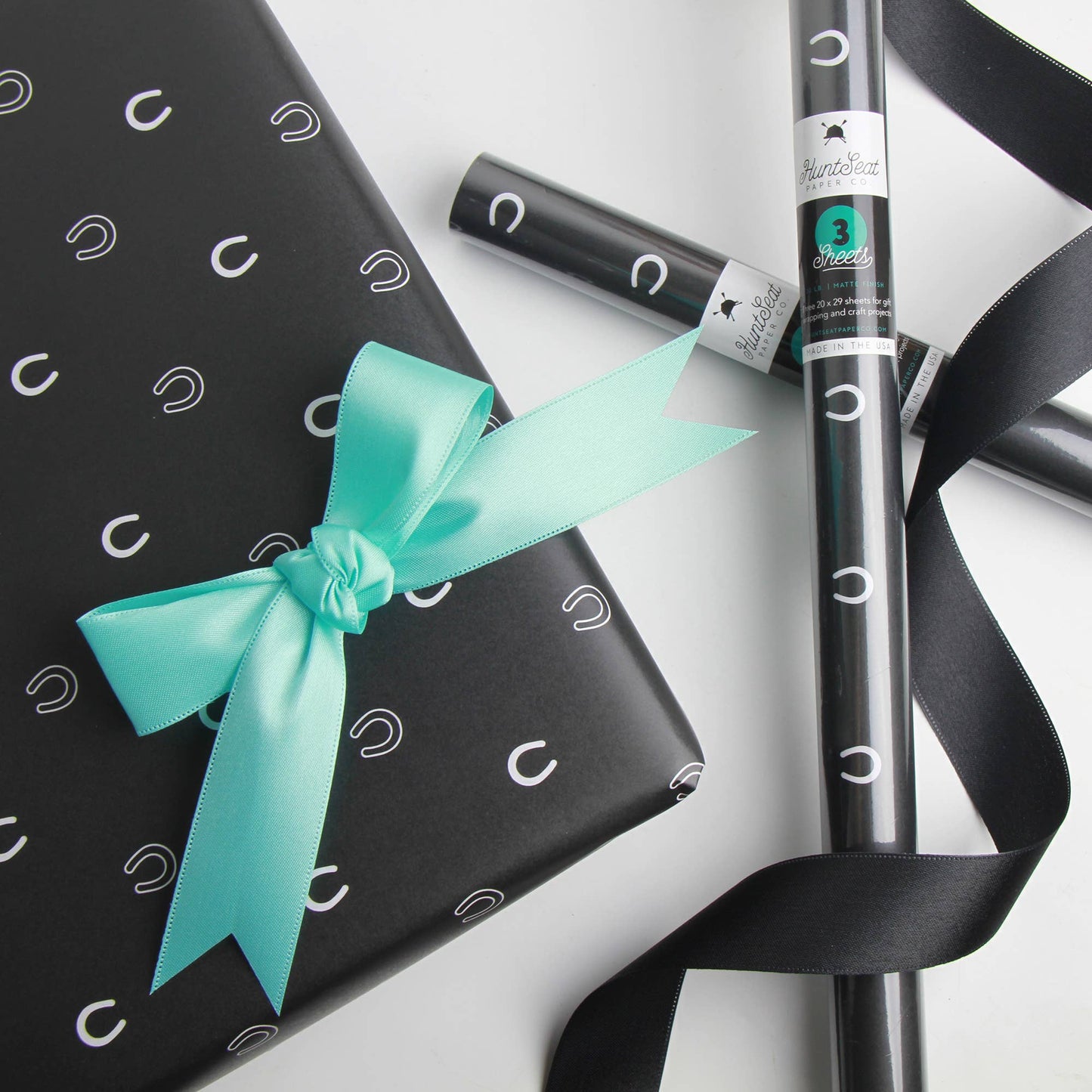 Black gift wrap with white horse shoe patterns, turquoise ribbon, and a roll of black ribbon on a light gray background.