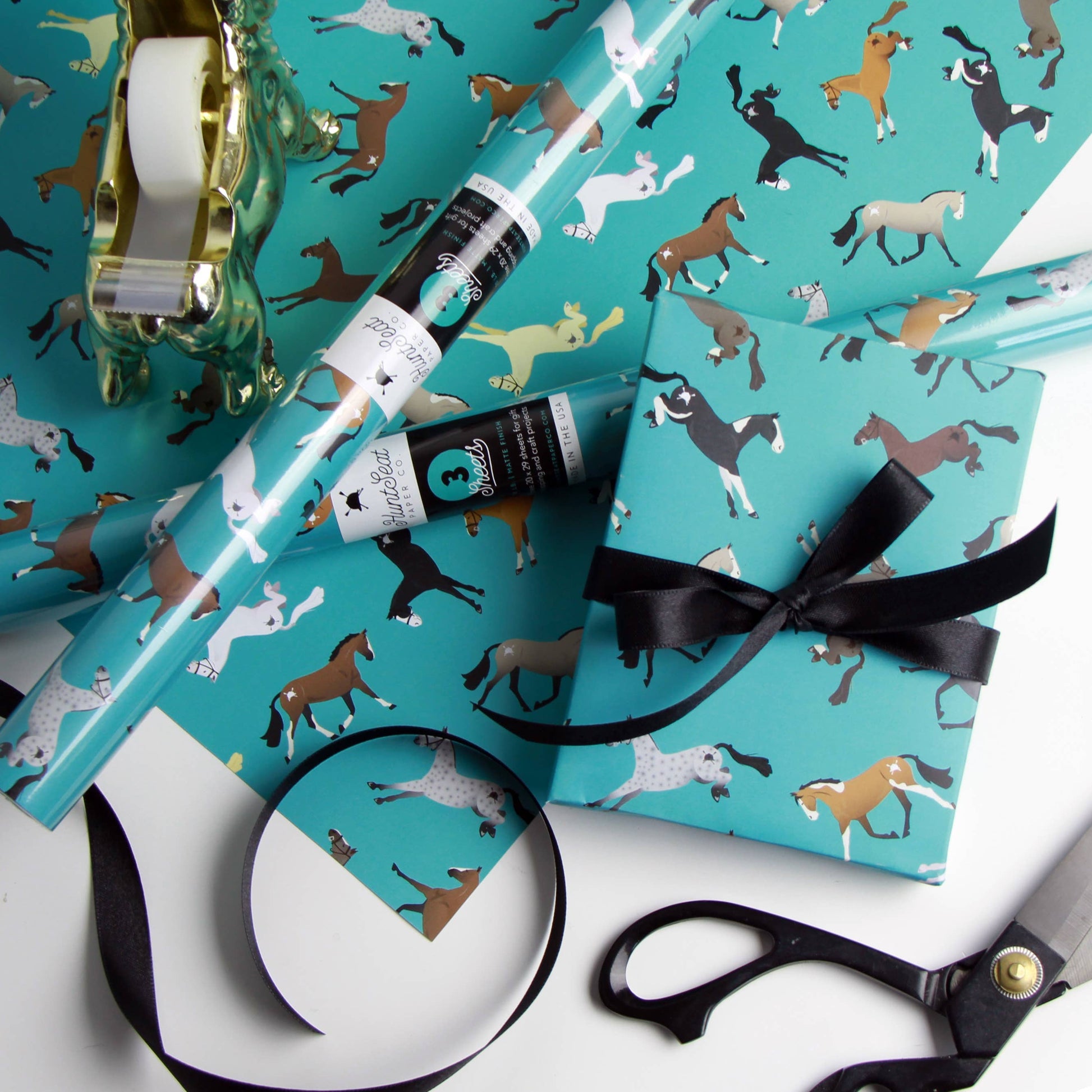 Turquoise wrapping paper with horse pattern, black ribbon, and scissors on a white background