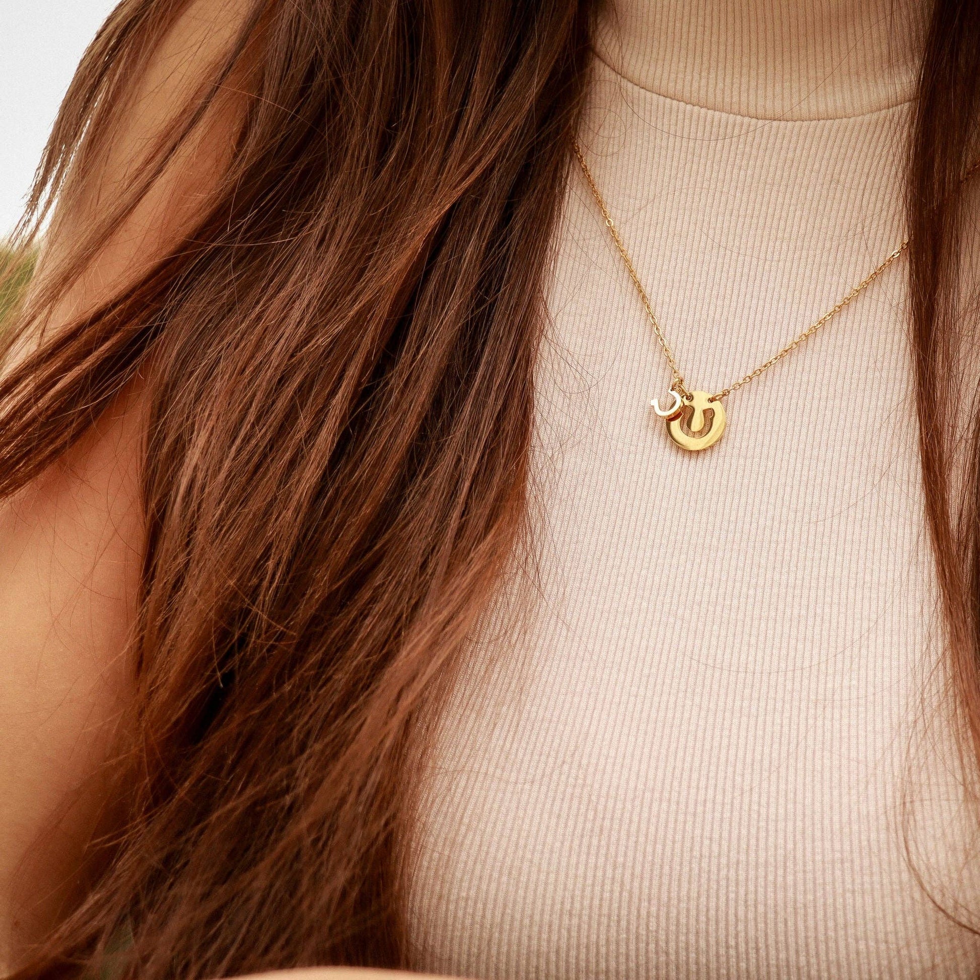 Gold necklace with horse shoe pendant worn by a person with long brown hair.