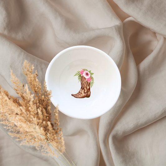 A white ceramic jewelry dish with a floral cowgirl boot design, placed on a beige fabric surface, accompanied by some dried plants.