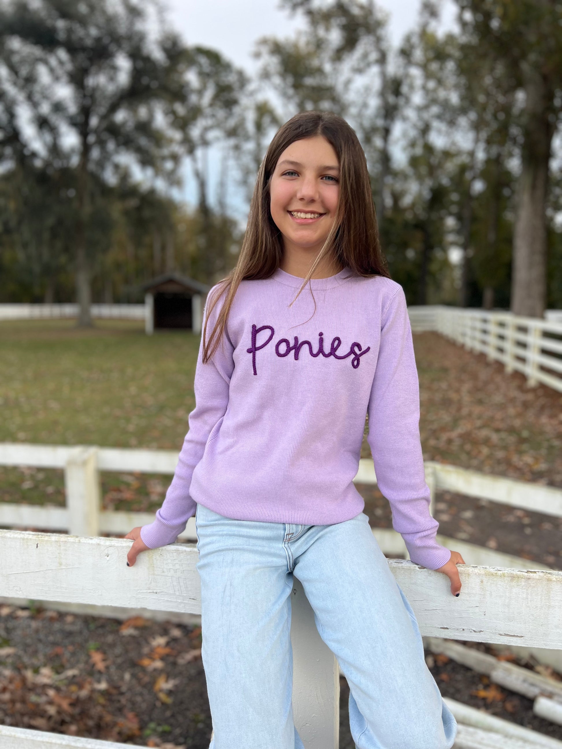 Teenage horseback rider wearing her favorite sweater that has ponies written across the front. She is sitting on a fence at a farm