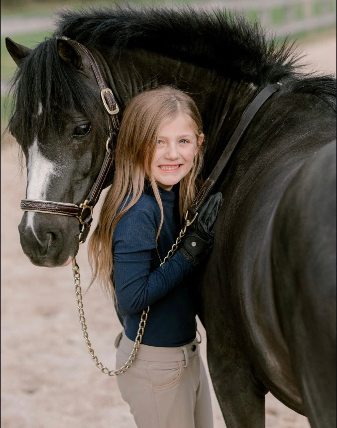 Girl standing next to a horse with a bridle on a dirt path, wearing a mesh mock neck riding top.