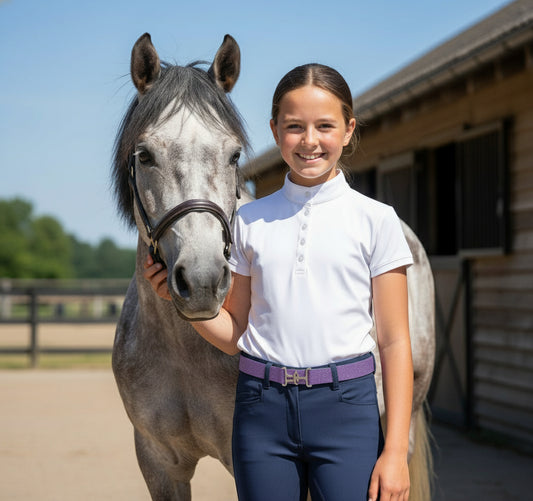 Girl in equestrian attire featuring a purple sparkle belt, standing next to a gray horse in an outdoor setting.