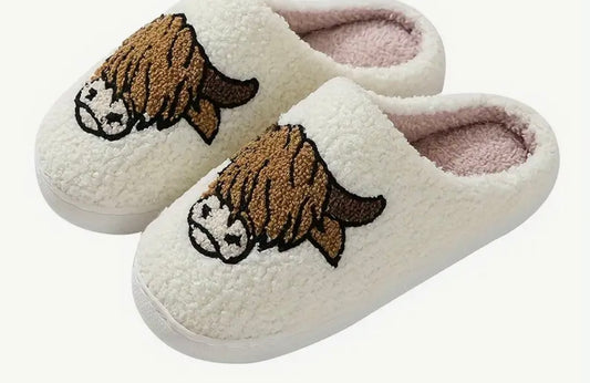 A pair of white slippers with a rubber sole and a decoration of a Highland cow on the top.