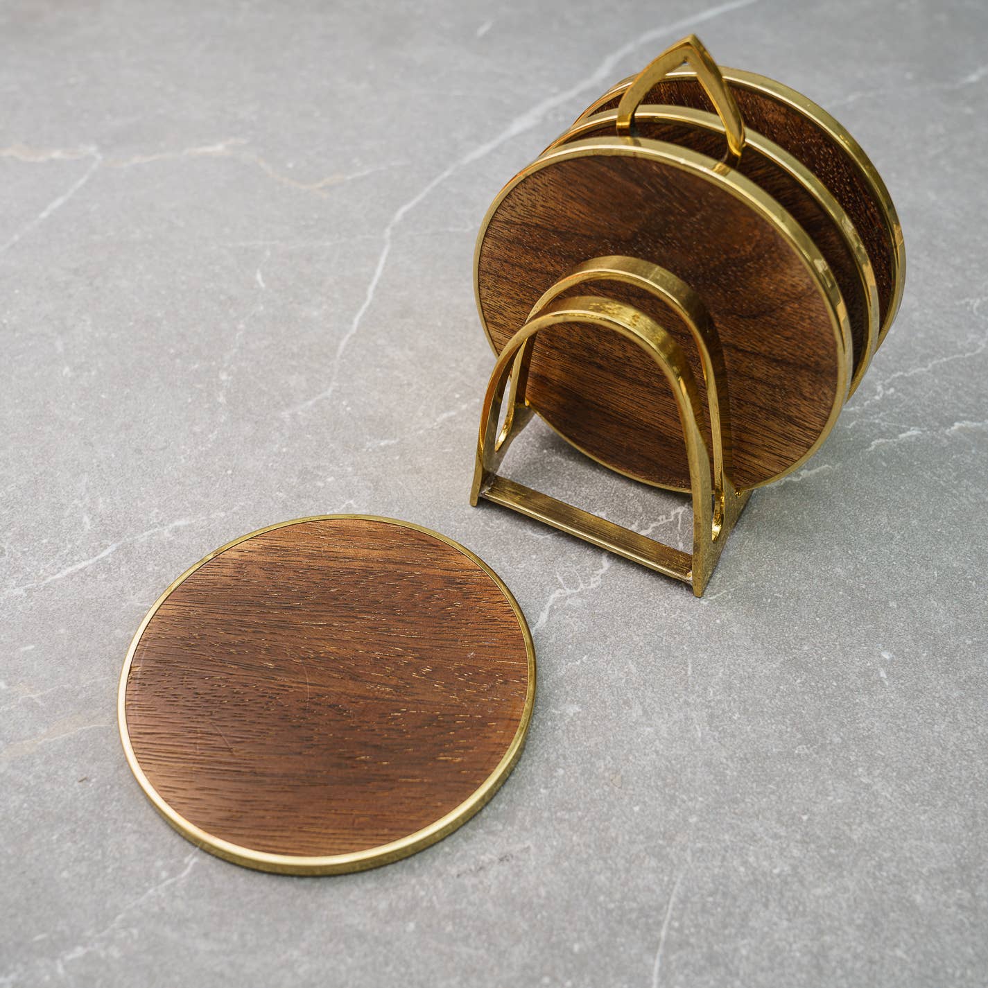 A set of four round wooden coasters with gold edges, accompanied by a brass stand, displayed on a grey surface.