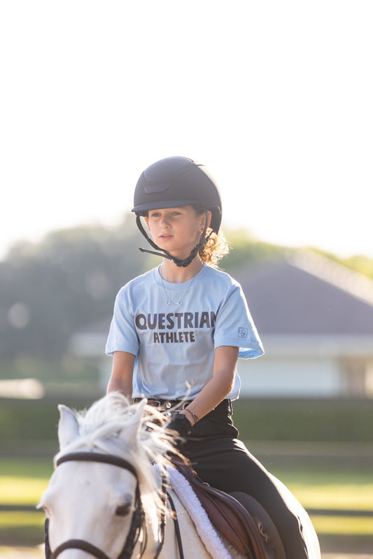 A child wearing a light blue crew neck t-shirt with 'EQUESTRIAN ATHLETE' printed on the front, sitting on a horse.