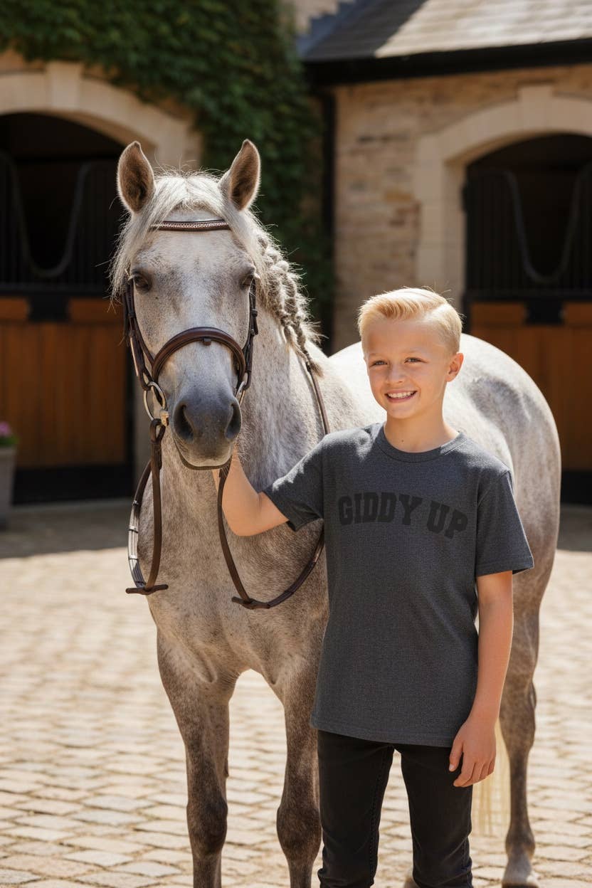 Young boy standing next to a gray horse in an equestrian setting wearing a gray "giddy up" t-shirt