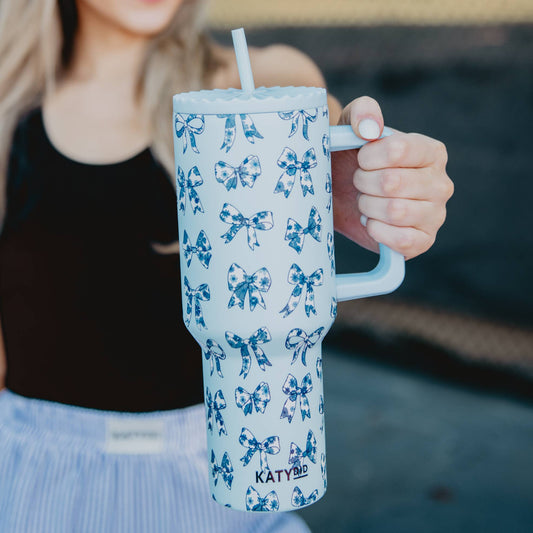 A person holding a blue tumbler with a floral pattern of blue ribbons and bows, featuring a reusable plastic straw and an adjustable straw cover.