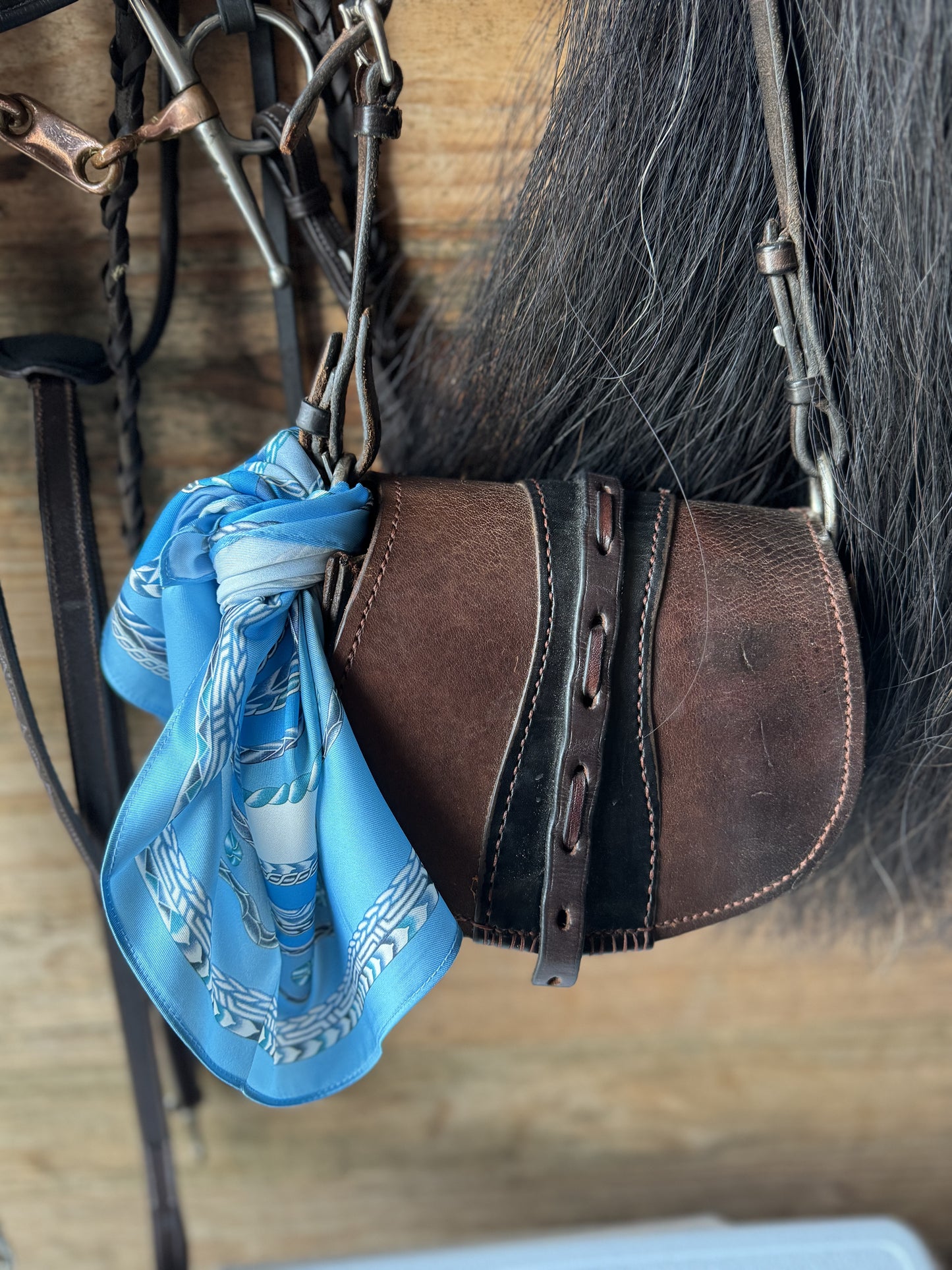 a blue silk with a horse head design tied to a crossbody leather purse, hanging with bridles next to a horse tail