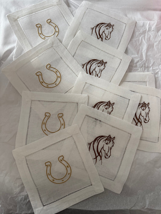 A collection of white linen cocktail napkins with an embroidered golden horseshoe design spread out on a surface.
