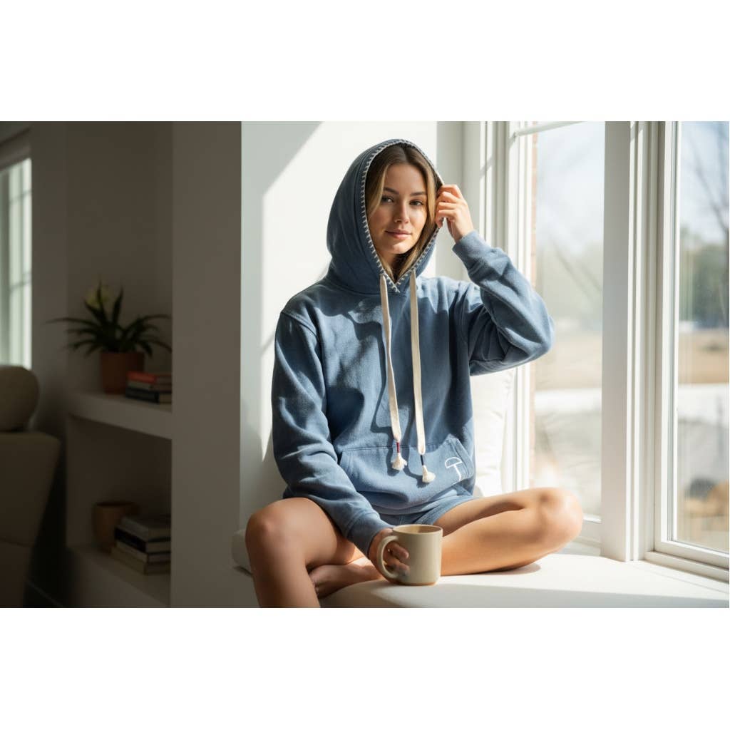 Person wearing a blue equestrian hoodie sitting by a window holding a mug.