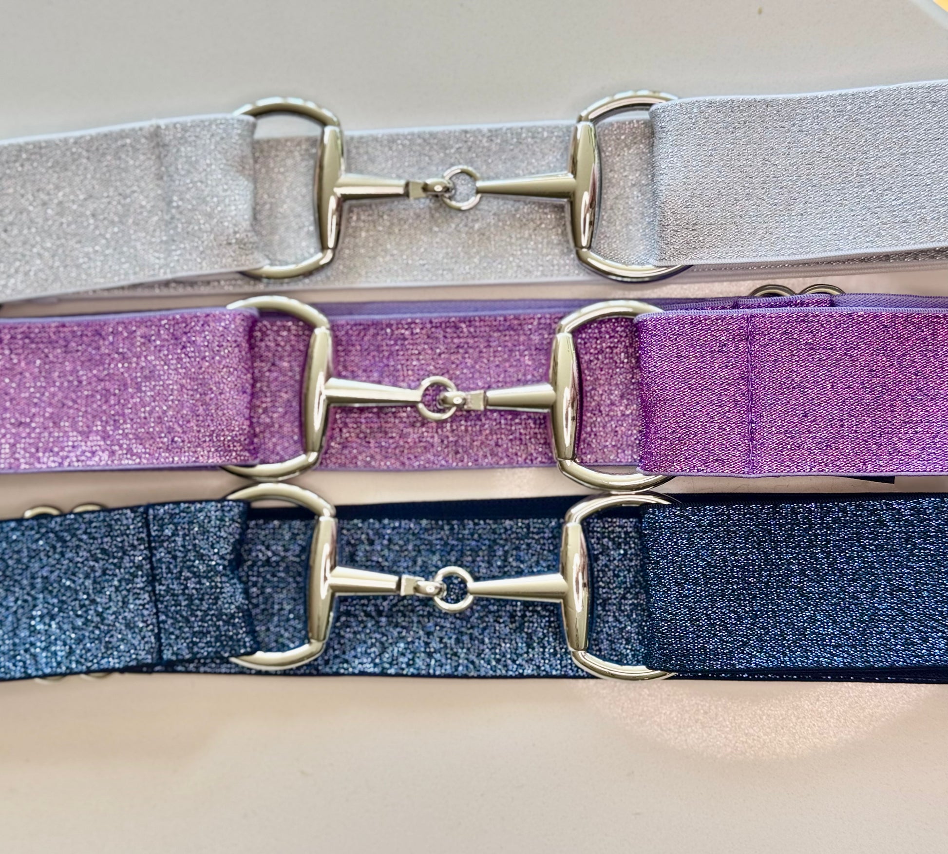 A collection of three sparkle bit belts in silver, purple, and blue colors with chrome hardware and a snaffle bit buckle closure.