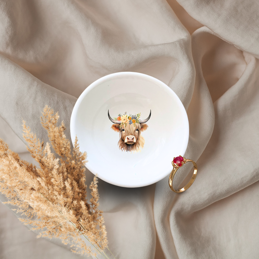 A white ceramic trinket dish with a illustration of a highland cow with a floral crown, displayed on a textured fabric with a ring and dried plants.