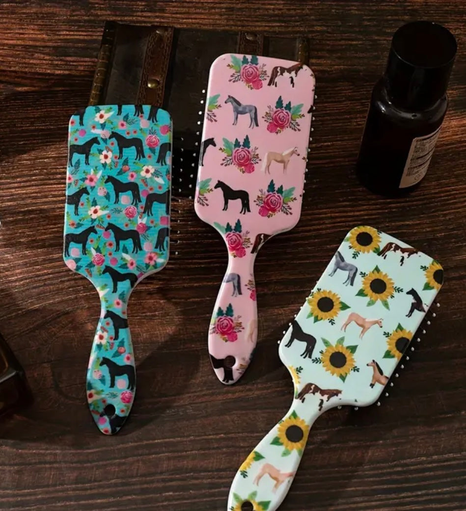 A set of three paddle brushes with colorful horse patterns, displayed on a wooden surface.