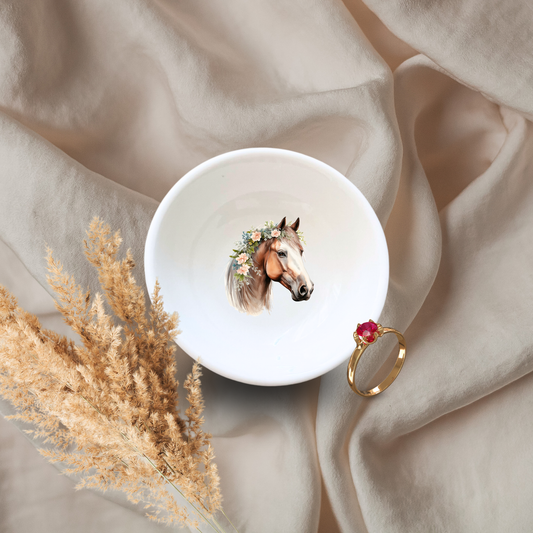 A white ceramic dish with a floral pattern and an illustration of a horse in the center, displayed on a fabric surface alongside some dried plants and a gold-colored ring.