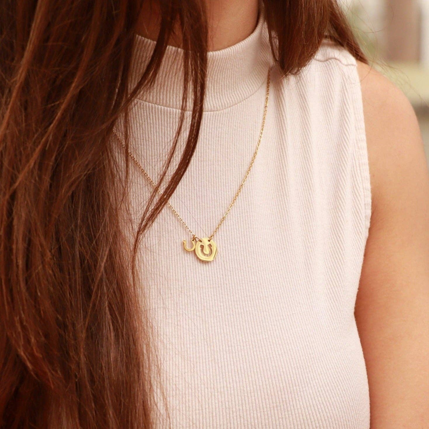 Person wearing a gold necklace with a heart and horseshoe pendant, blurred background