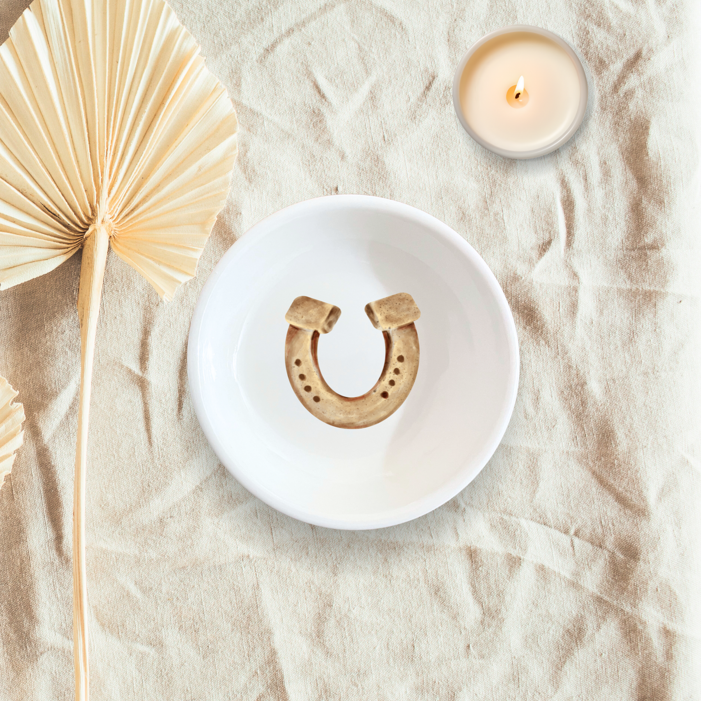 A white handmade ceramic dish with a horsehoe design in the center, displayed on a beige textile surface alongside a candle and a dried palm leaf.