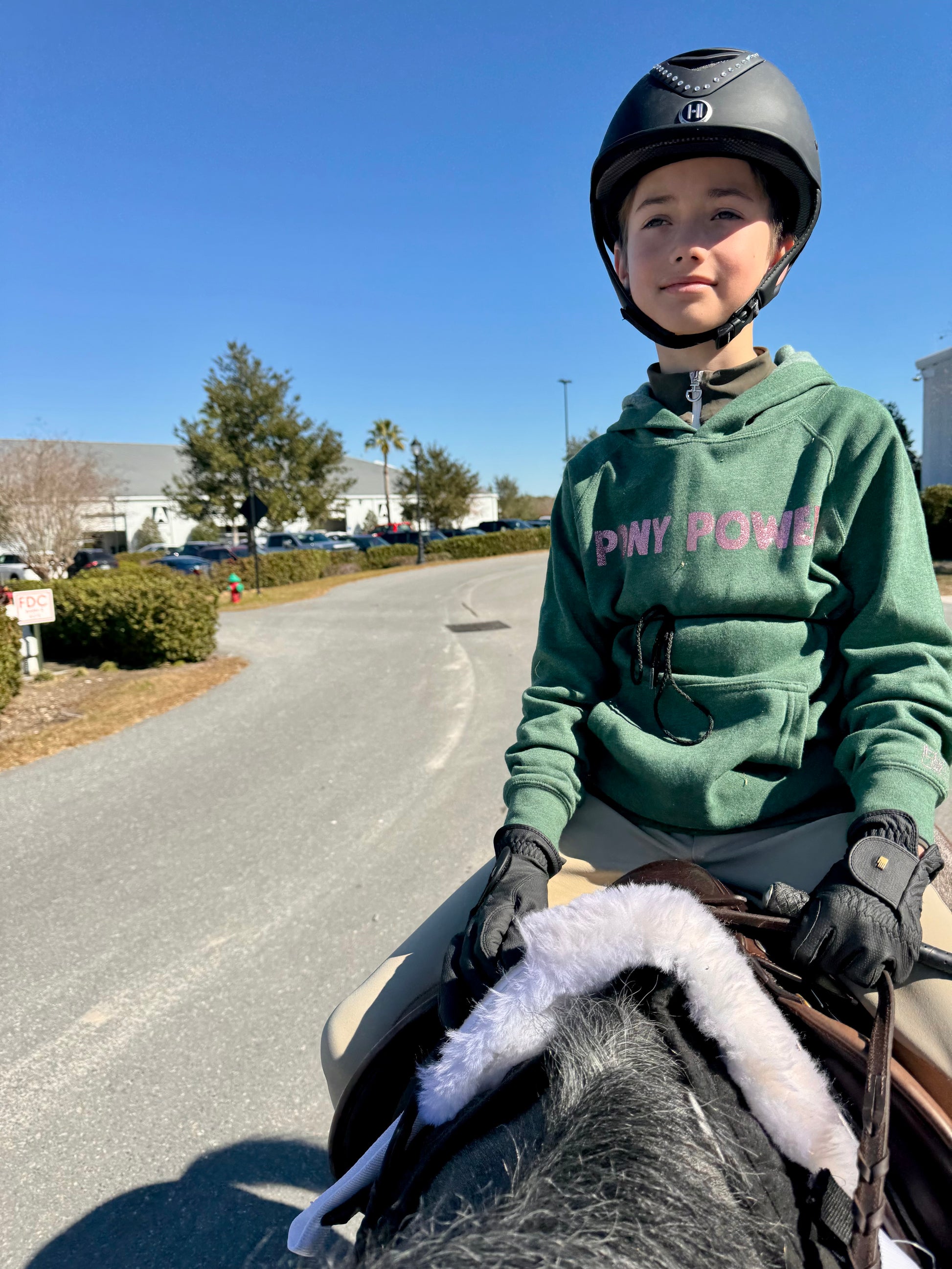 boy riding a pony wearing his green "pony power" hoodie, outside on a sunny day