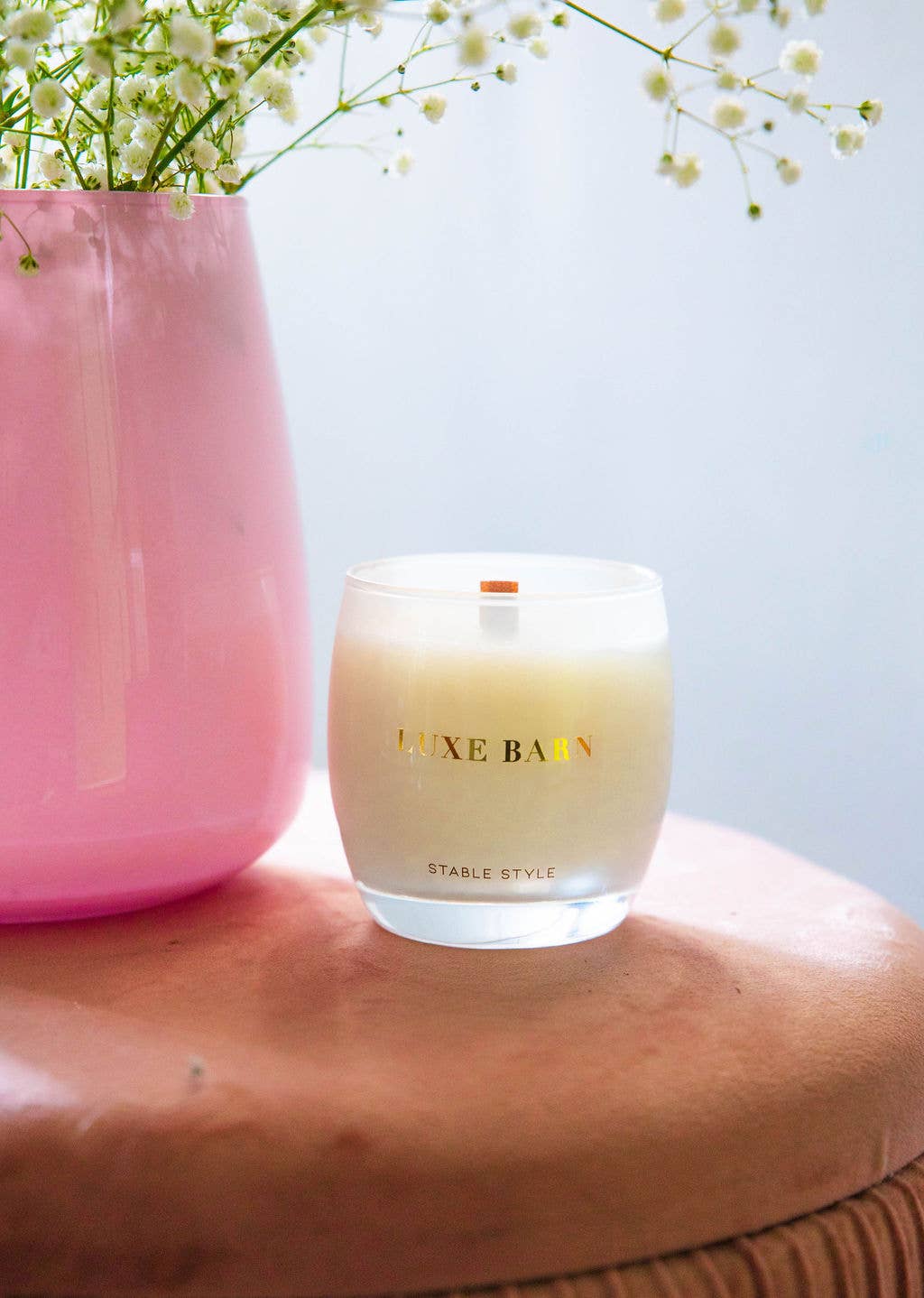 A soy wax candle with a wooden wick, in an elegant white jar with delicate gold font. The candle is placed on a pink surface, with a vase of flowers in the background.