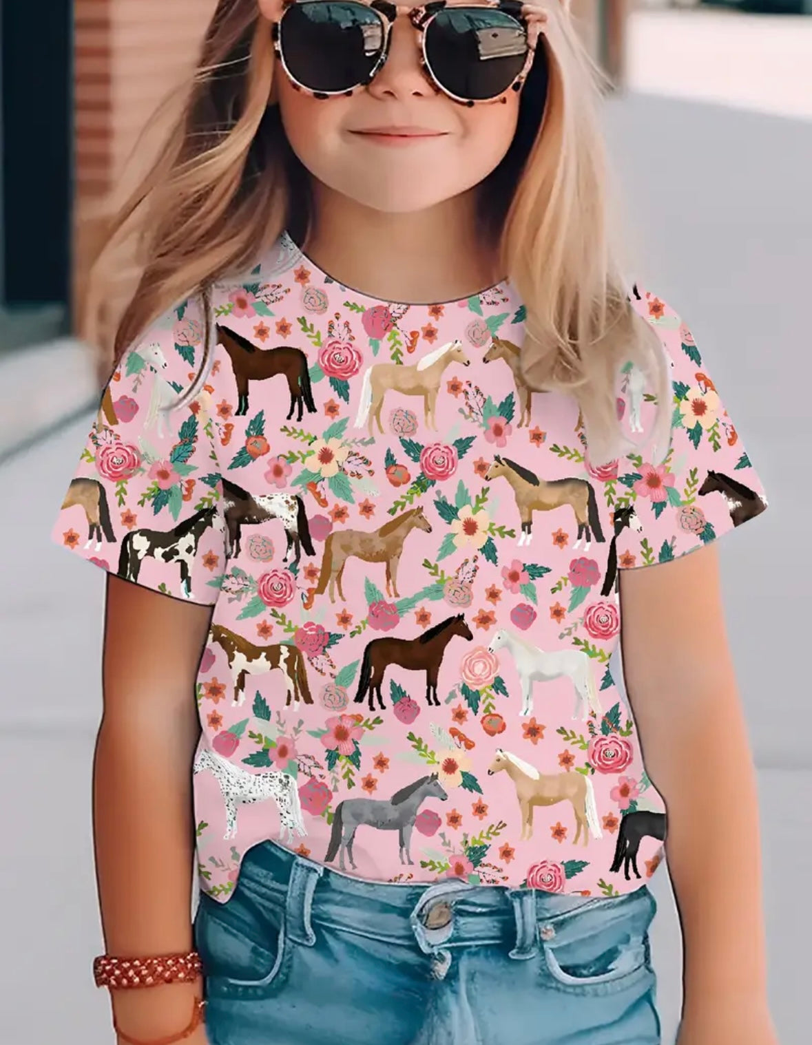 A child wearing sunglasses and a pink crewneck t-shirt with a colorful horse pattern.